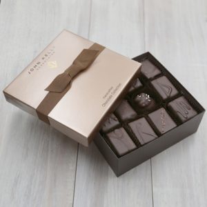 A partially opened box of the 24 Piece Signature Handcrafted Chocolate Collection. Some of the chocolates are visible. The lid is sitting partially on the box. The lid is a light brown color and has a brown ribbon on it.