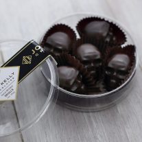 An open container with five dark chocolate skulls in it. The lid of the contained is leaning against the container. The container is a clear, plastic, round container with a black John Kelly Chocolates logo